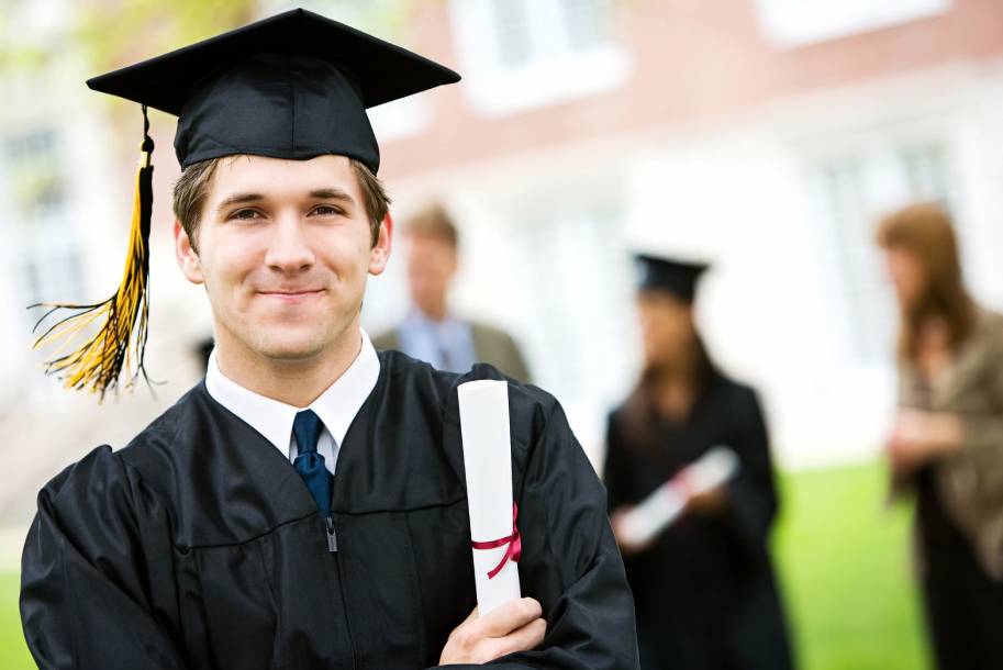 Graduate student holding his diploma looking at the camera smiling without showing his teeth.